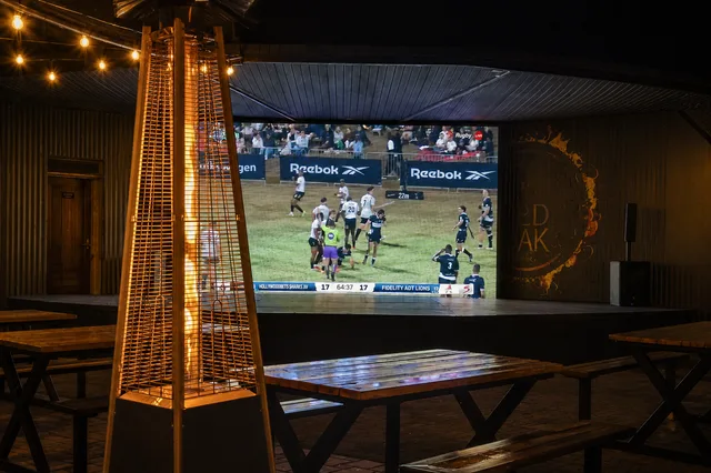 Rugby on the big screen with a patio heater and empty wooden tables under warm string lights.