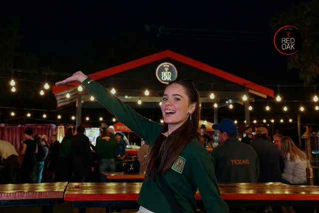 A woman in a Springbok jersey raising her arm with the Red Oak building and string lights behind her.